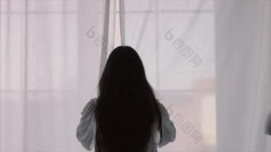 长发黑发女孩打开窗帘窗口拉伸在早晨自然光4k后视图。性感的女人站在举起手臂，在室内<strong>剪影</strong>4k。自由<strong>女性</strong>时尚服装